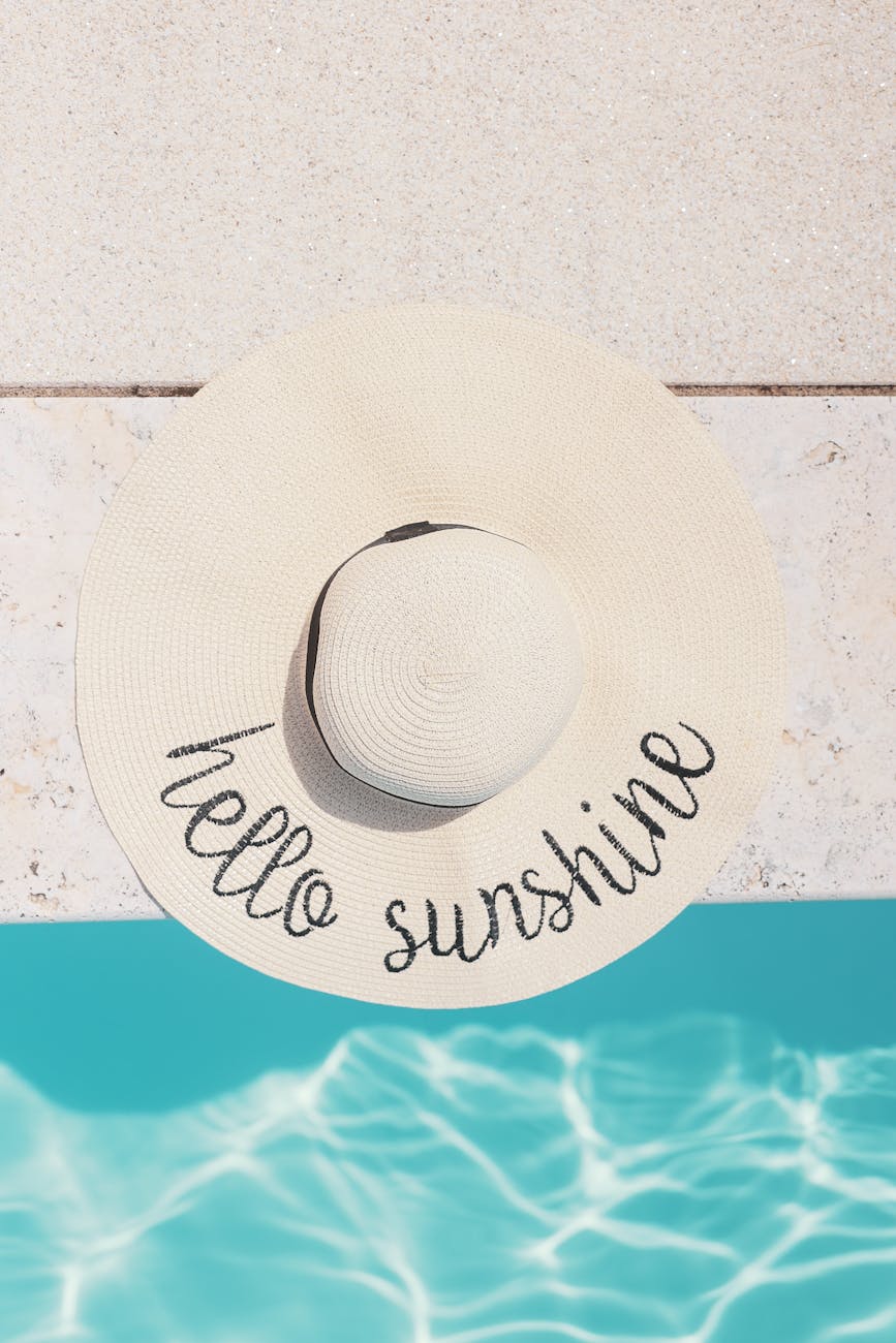 A stylish beach hat with 'Hello Sunshine' text sits by a clear blue pool.