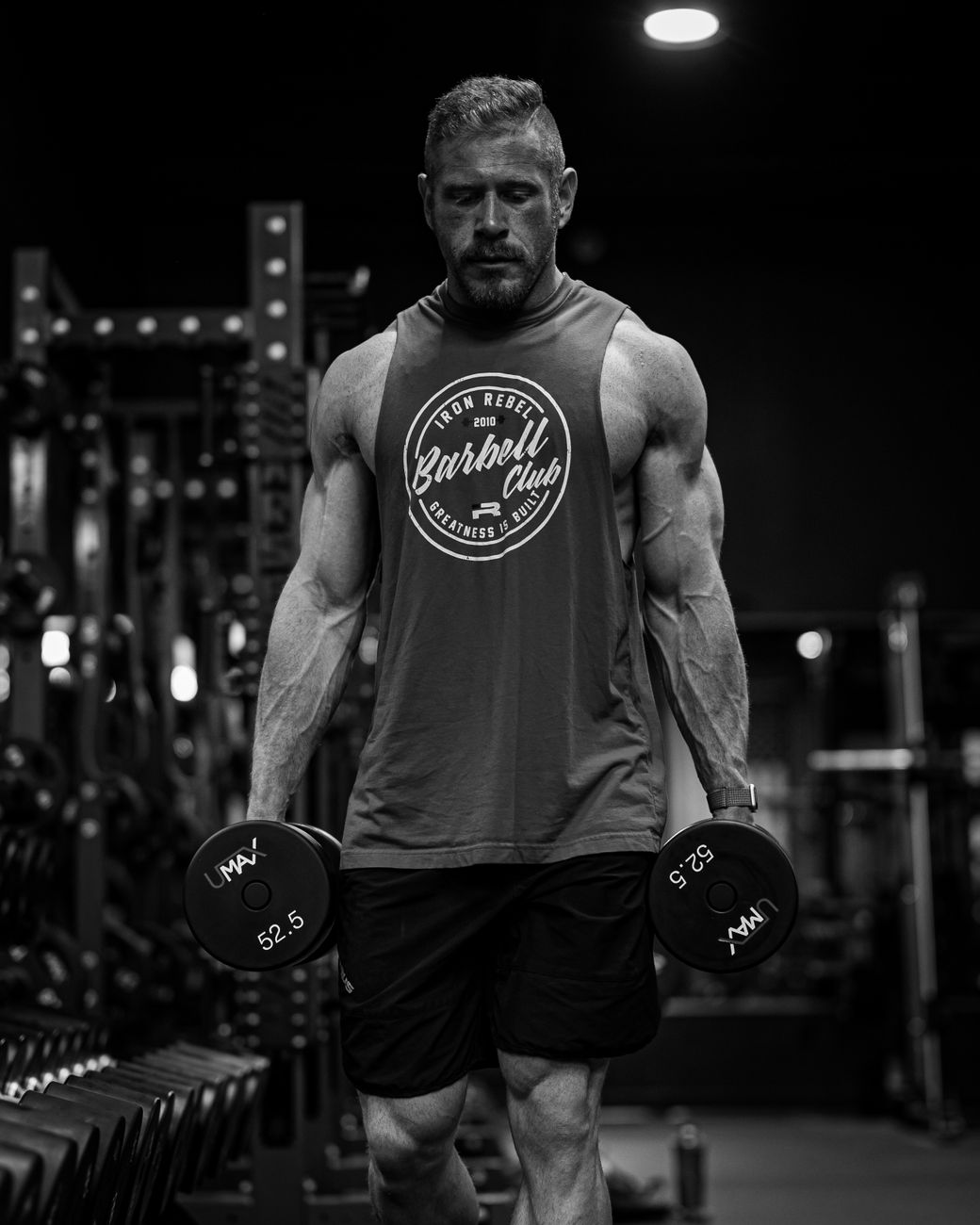 Black and white photo of a muscular man lifting dumbbells in a gym, showcasing strength and focus.