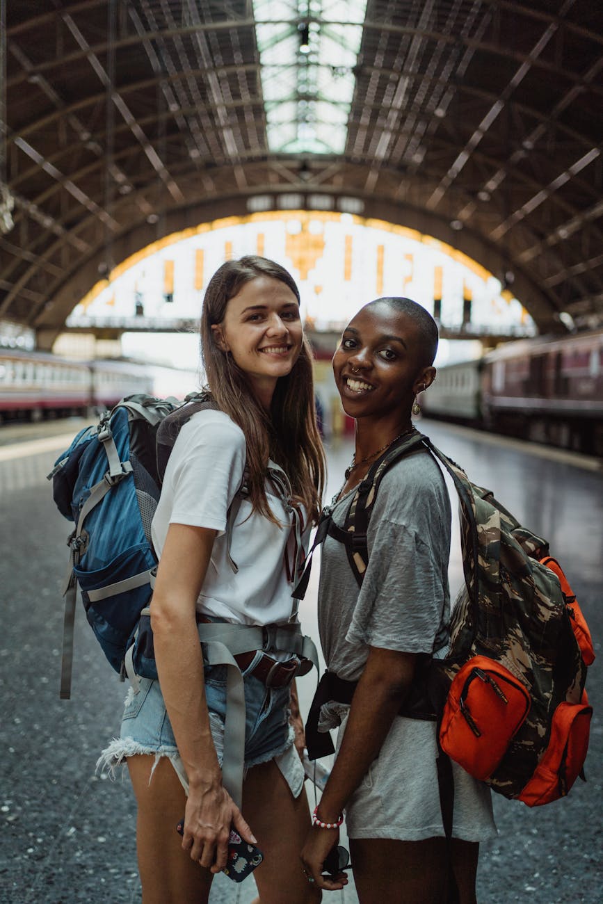 Two cheerful women backpackers at a vintage railway station, ready for adventure.
