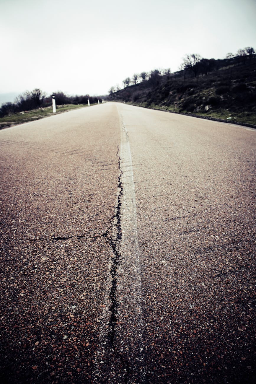 A cracked rural asphalt road leading through a scenic landscape under a cloudy sky.