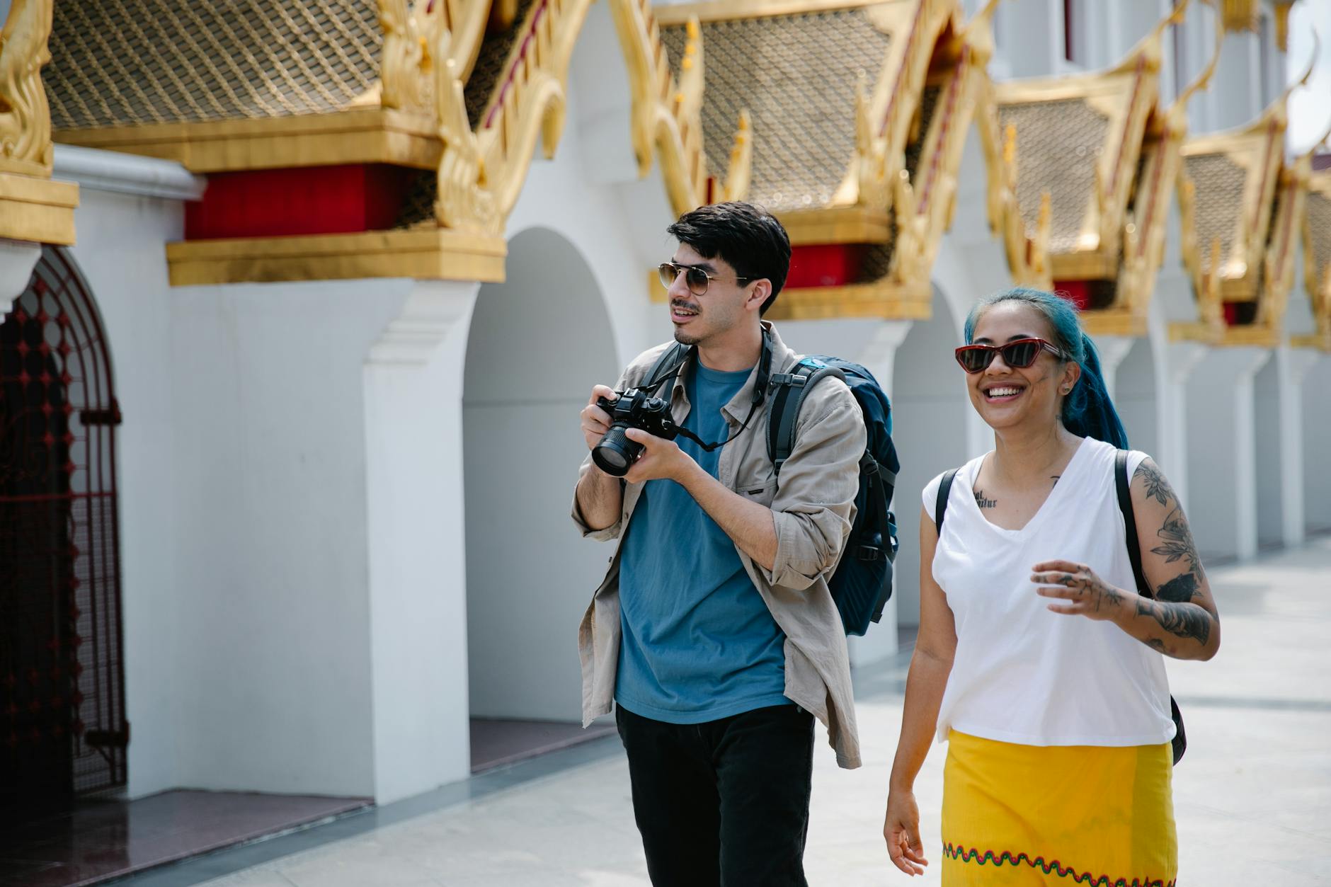 Two tourists with backpacks and cameras enjoy a sunny day exploring ornate Thai architecture.