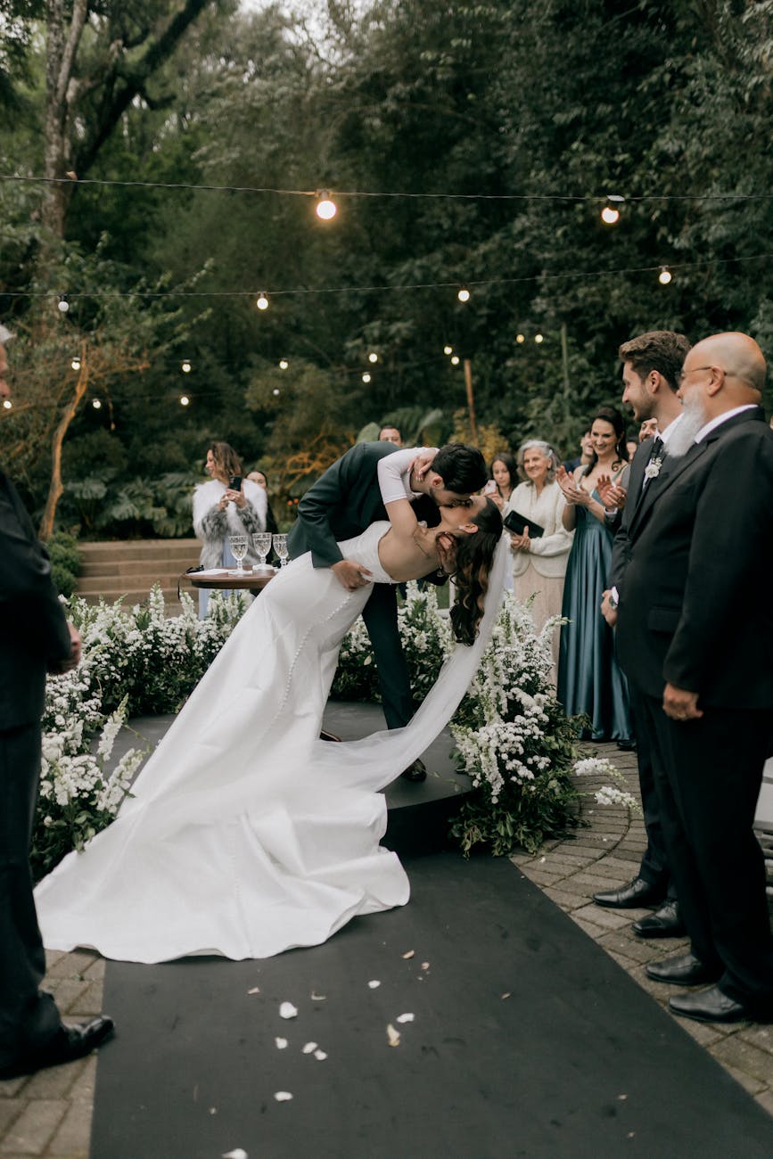 A couple's romantic kiss at an outdoor wedding ceremony surrounded by guests and flowers.