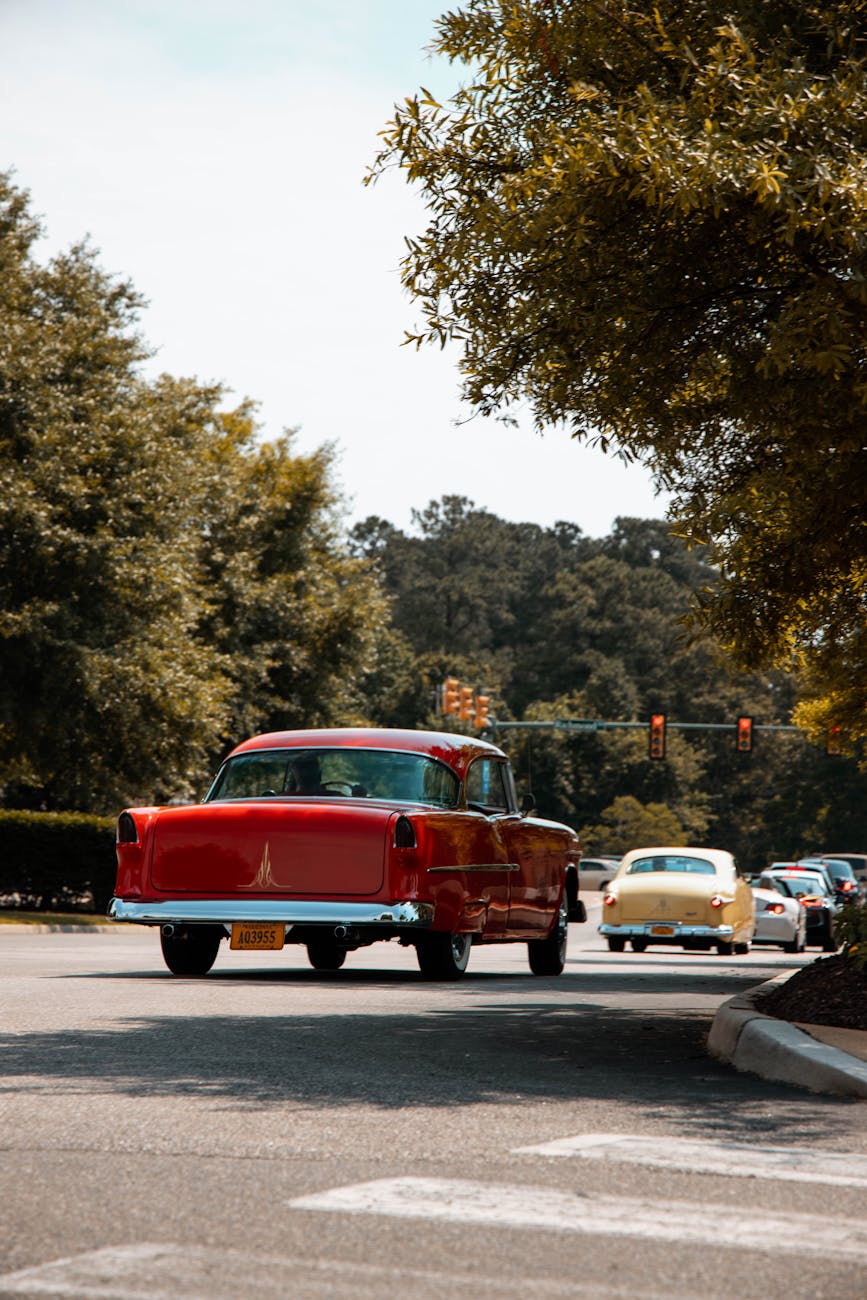 Retro red and yellow cars on a leafy urban road under bright skies.