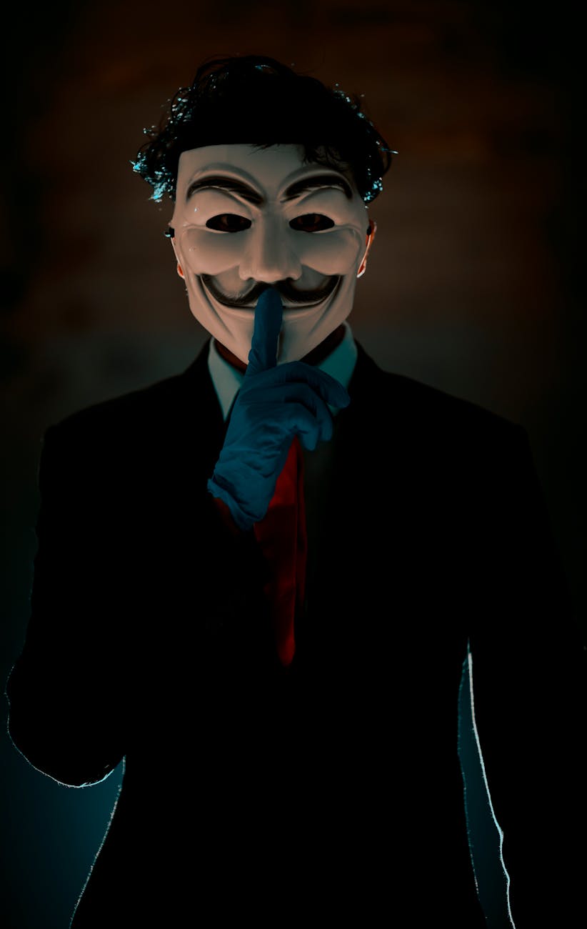 A person in a suit with a mask holding a finger to their lips in a shadowy atmosphere.