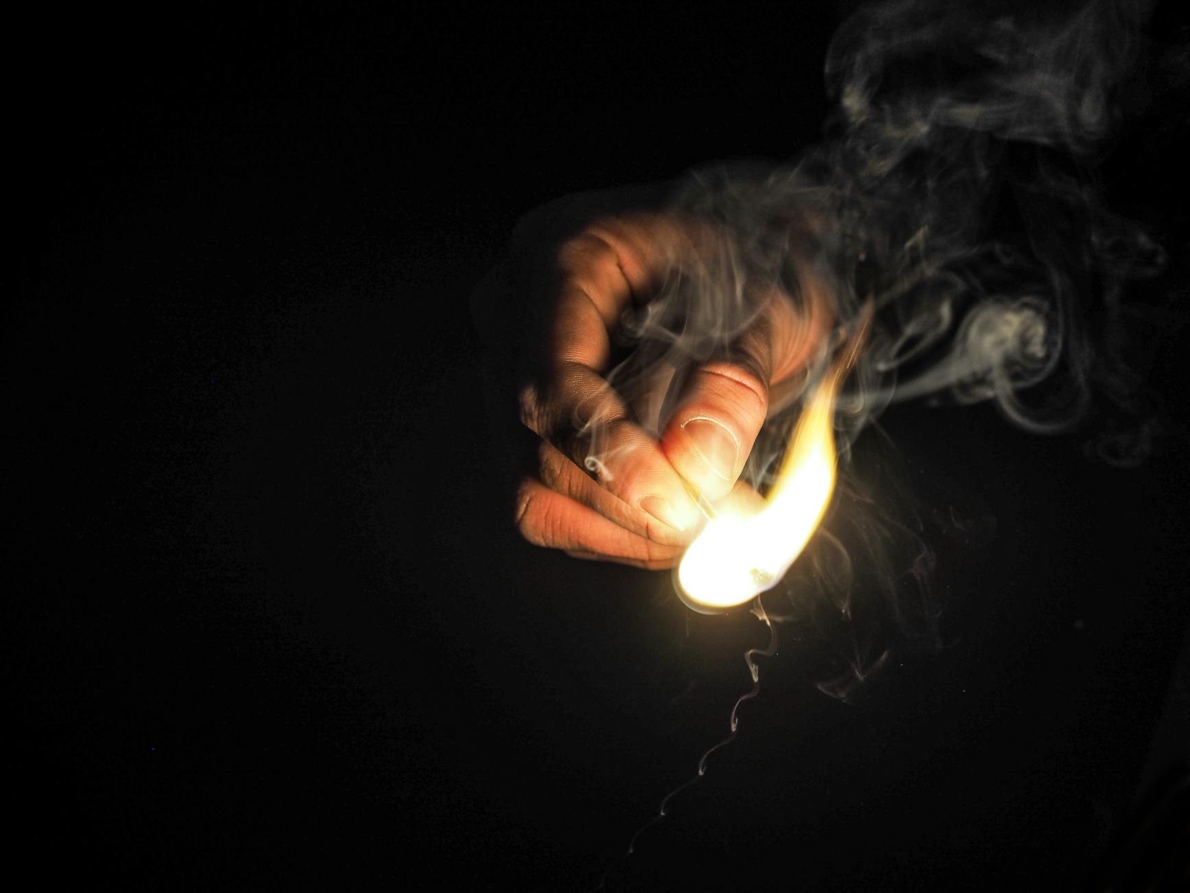 A close-up of a hand holding a bright flame with smoke against a dark background.
