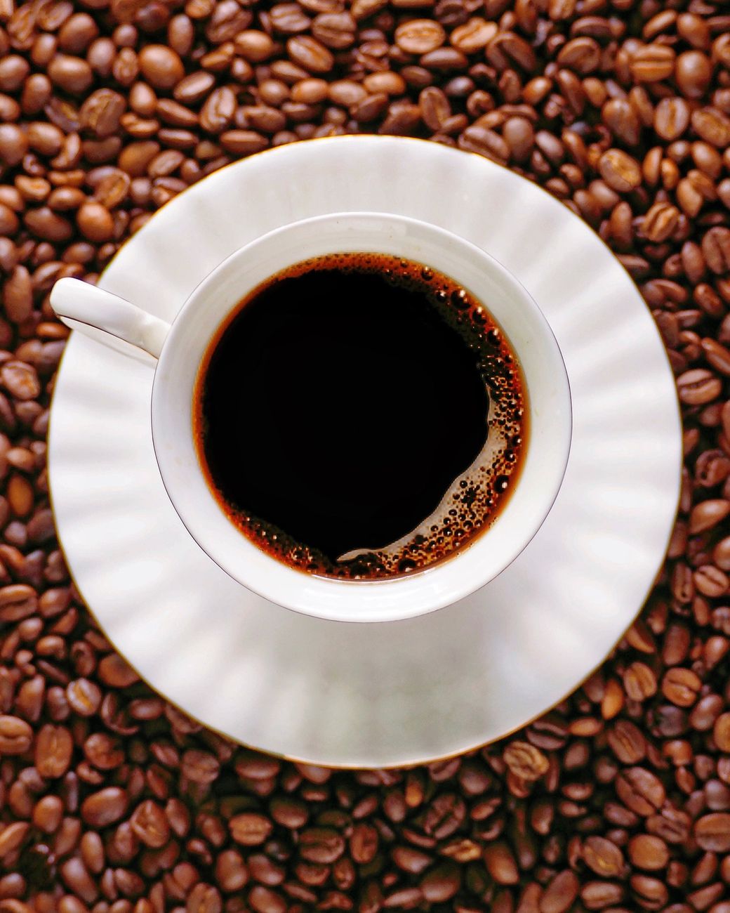 A white cup filled with coffee placed on roasted coffee beans, viewed from above.