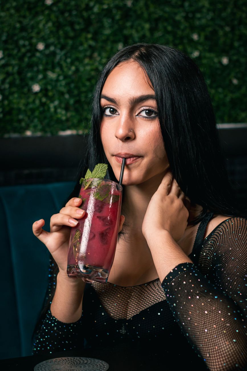 A woman sips a vibrant cocktail with mint garnish in a trendy indoor setting.