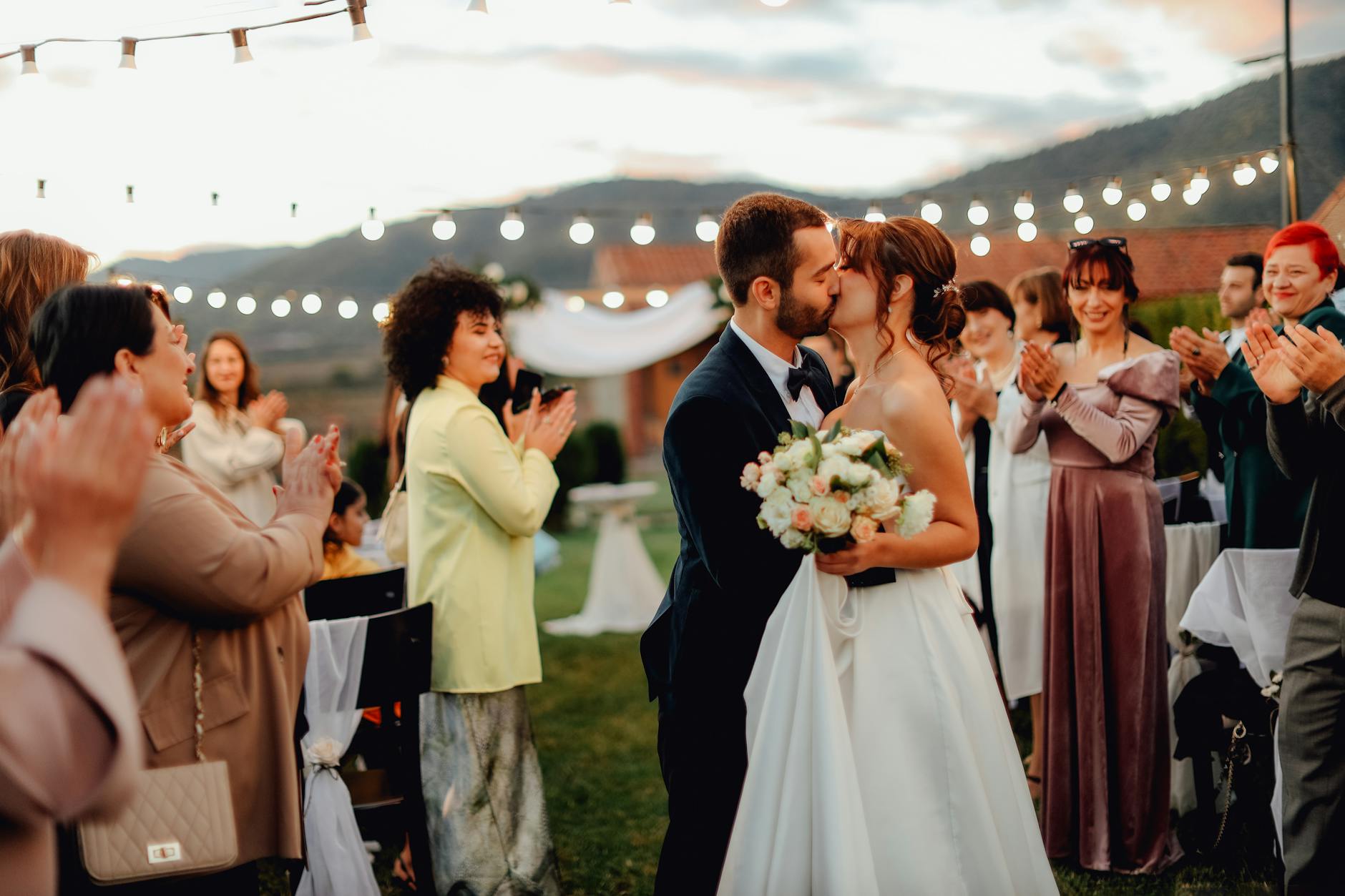 A bride and groom share a passionate kiss outdoors with guests applauding under string lights.