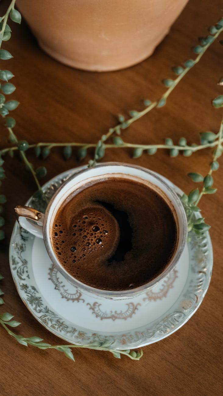 A close-up of Turkish coffee in a decorative cup with a plant on a wooden table.
