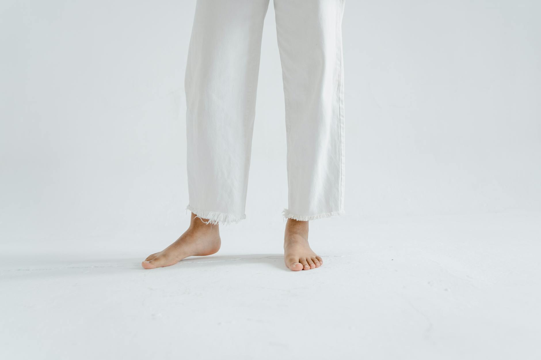 Close-up of barefoot legs in white pants against a minimalist white background.