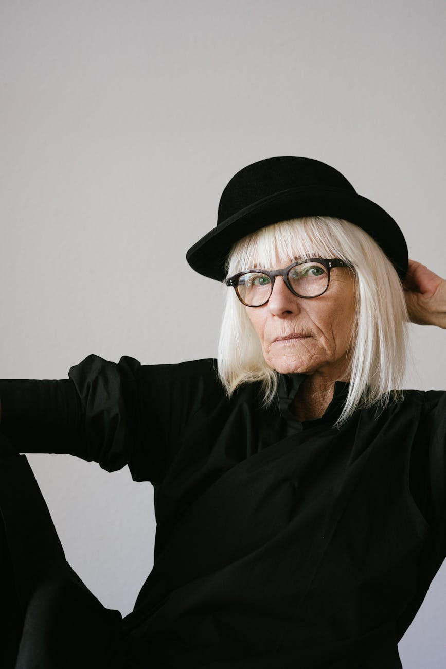 A senior woman in a black hat and glasses, exuding elegance and wisdom, against a neutral backdrop.