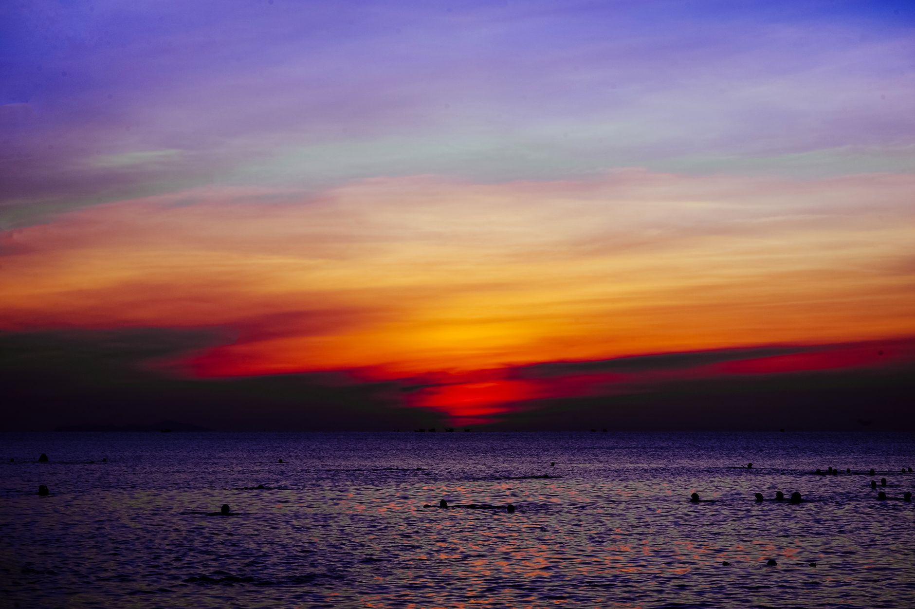 Vibrant sunset casting colorful hues over a serene ocean with silhouetted swimmers.