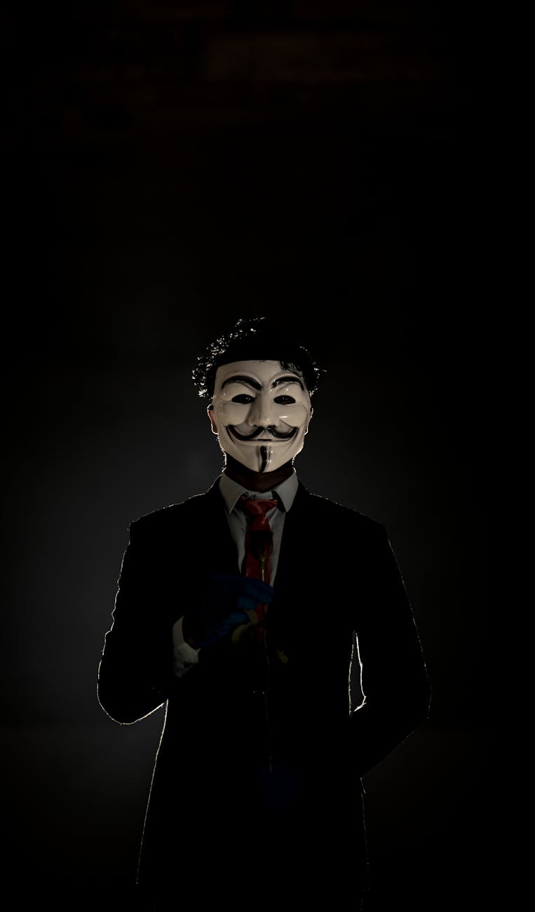 Mysterious figure in a suit wearing an anonymous mask in a shadowy environment.