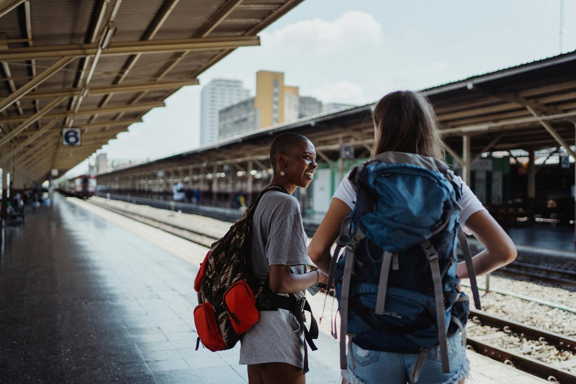Two diverse female backpackers wait on a sunny railway platform, ready for adventure.