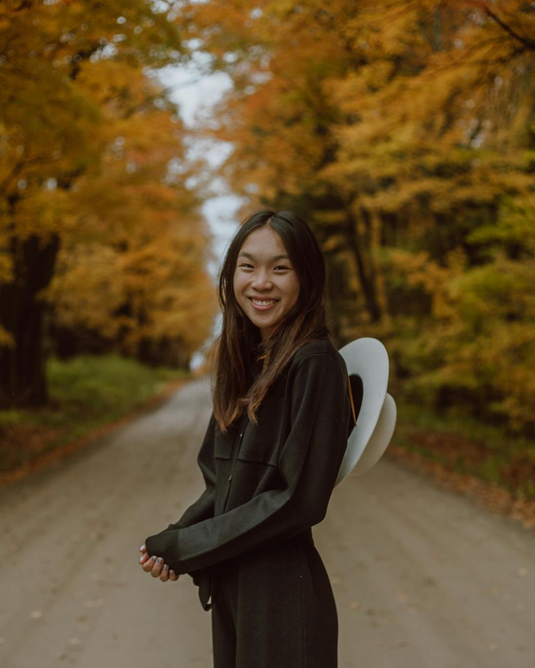 A cheerful woman in a hat and jacket walking along a dirt road lined with vibrant autumn foliage.
