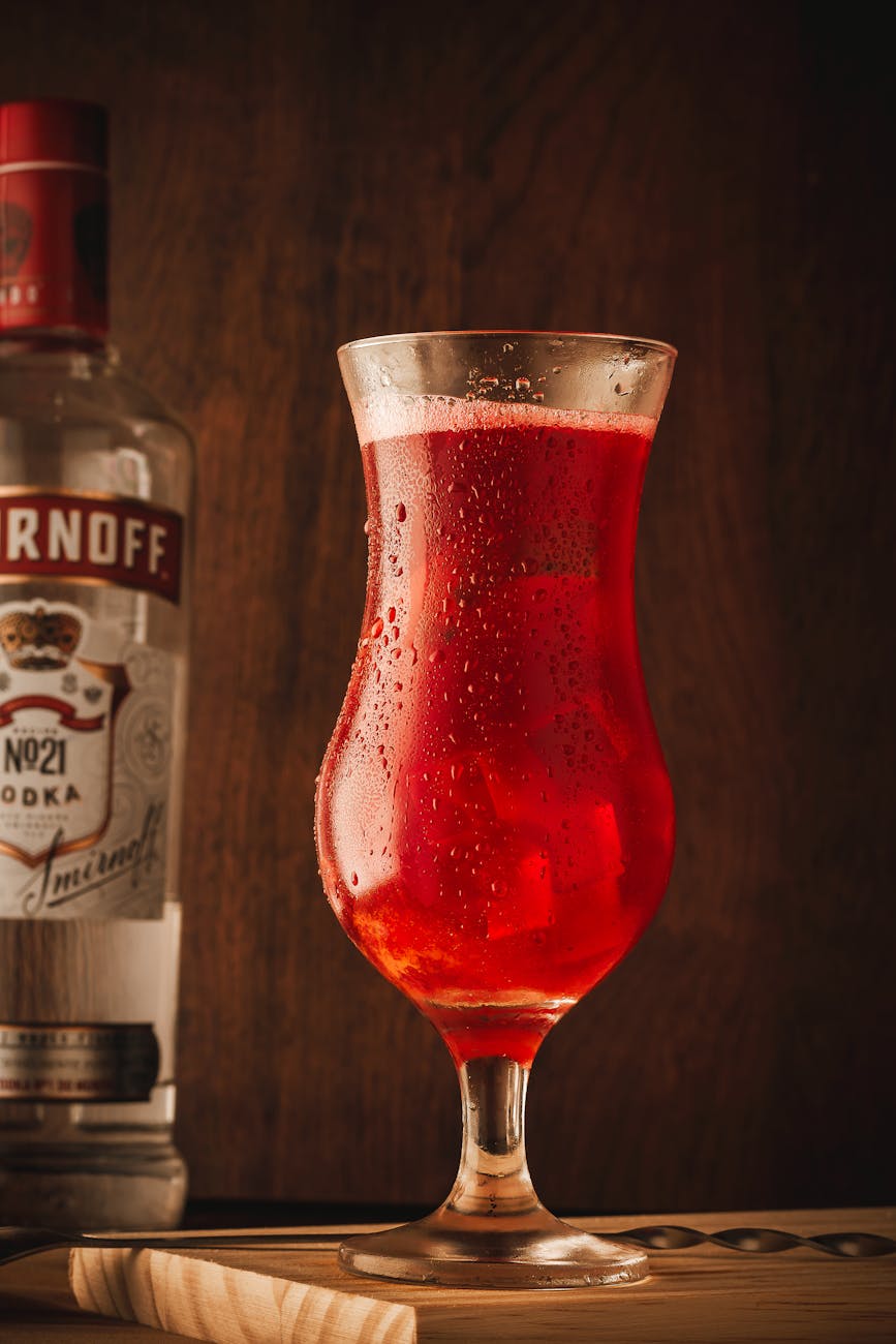Chilled red cocktail in hurricane glass beside Smirnoff vodka bottle on a wooden table.