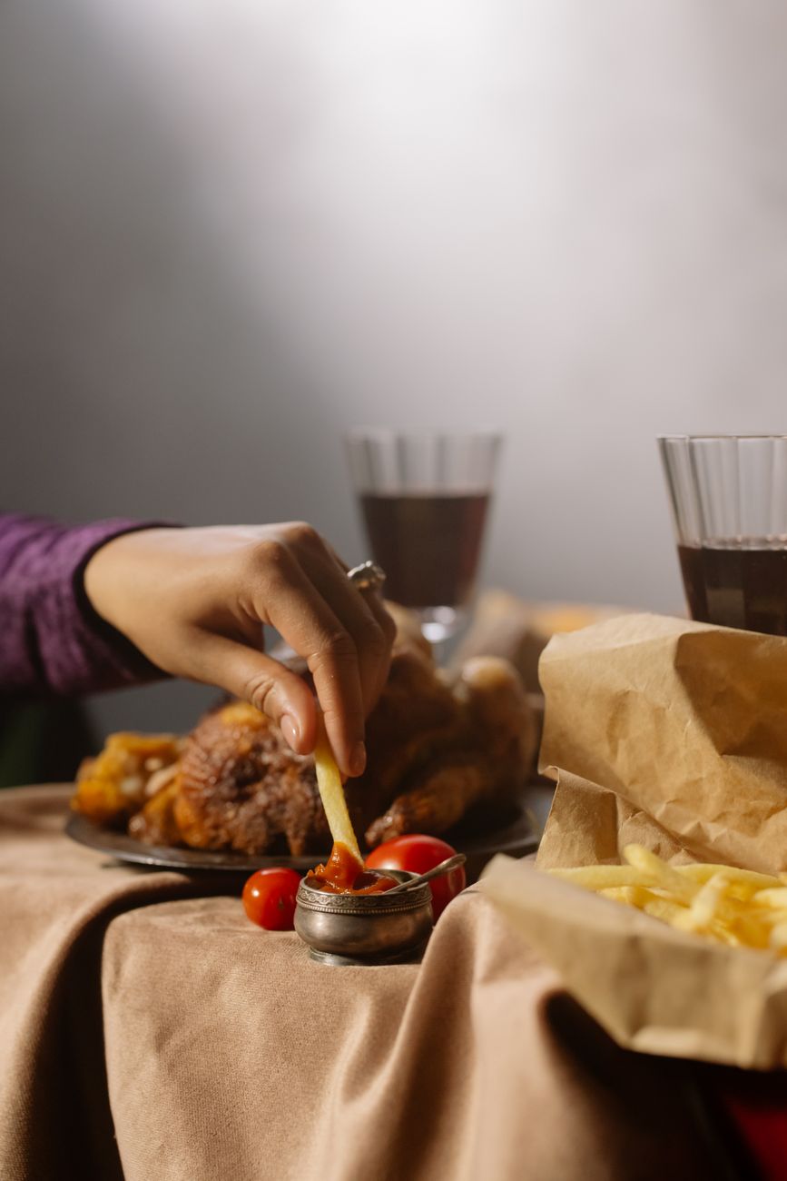 A hand dips a fry into ketchup, surrounded by fries and meat dishes, with glasses of wine in a cozy setting.