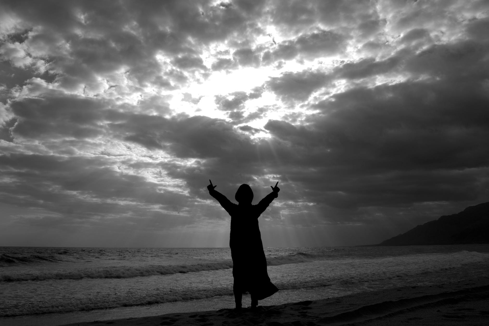 A person stands in silhouette on As Sifah Beach, Oman, beneath dramatic clouds and crepuscular rays.
