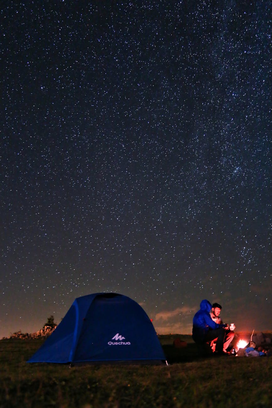 A serene night camping scene with a tent and people by a campfire under a starry sky.