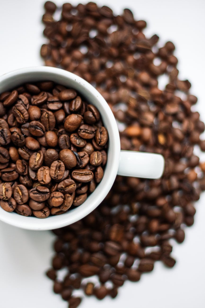 A top-down view of roasted coffee beans overflowing in a white mug on a white surface.