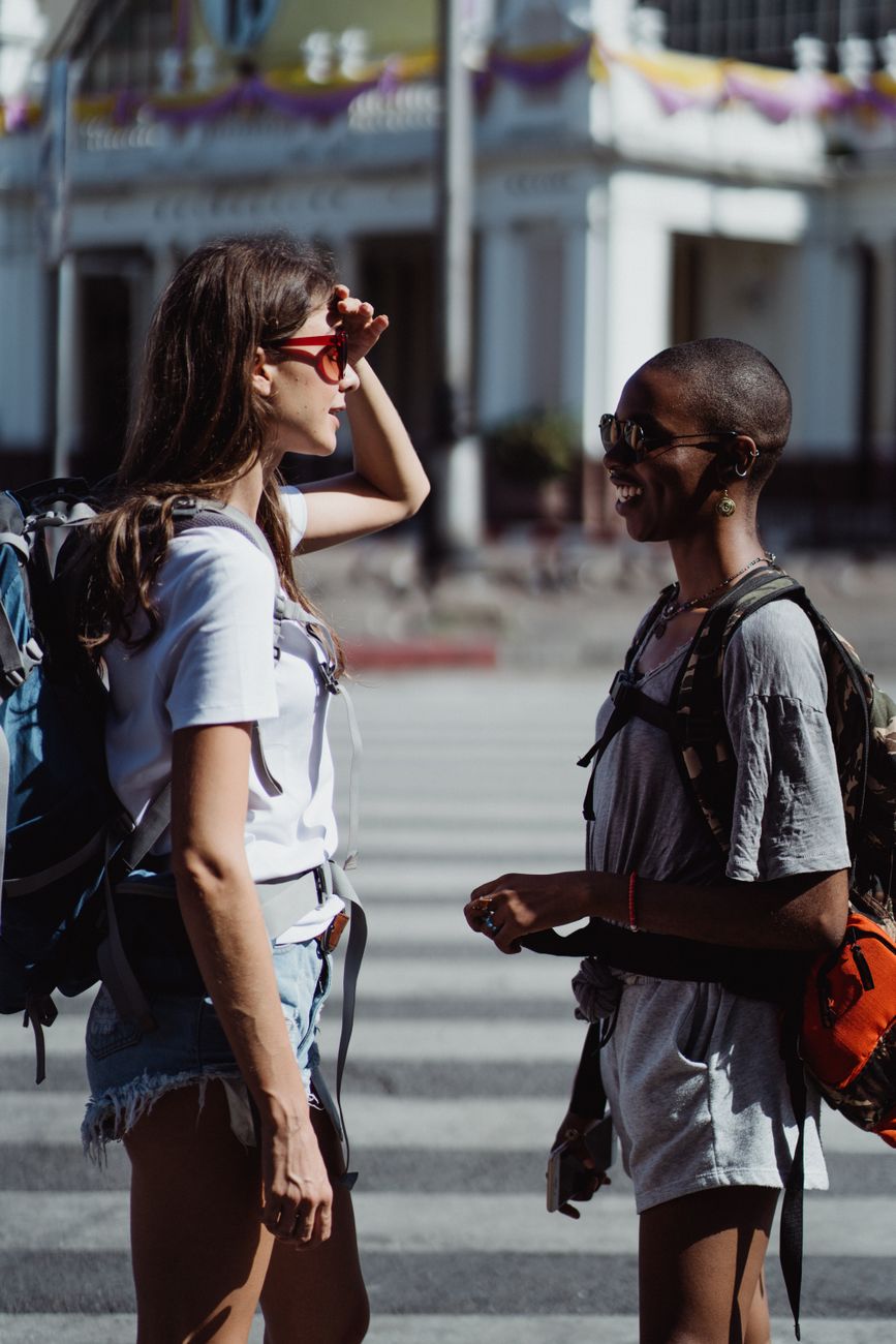 Two women with backpacks conversing on a sunny day, showcasing travel camaraderie.