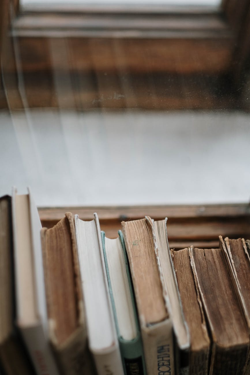 Close-up of vintage, worn-out books lined up on a wooden shelf by a window, creating a cozy feel.