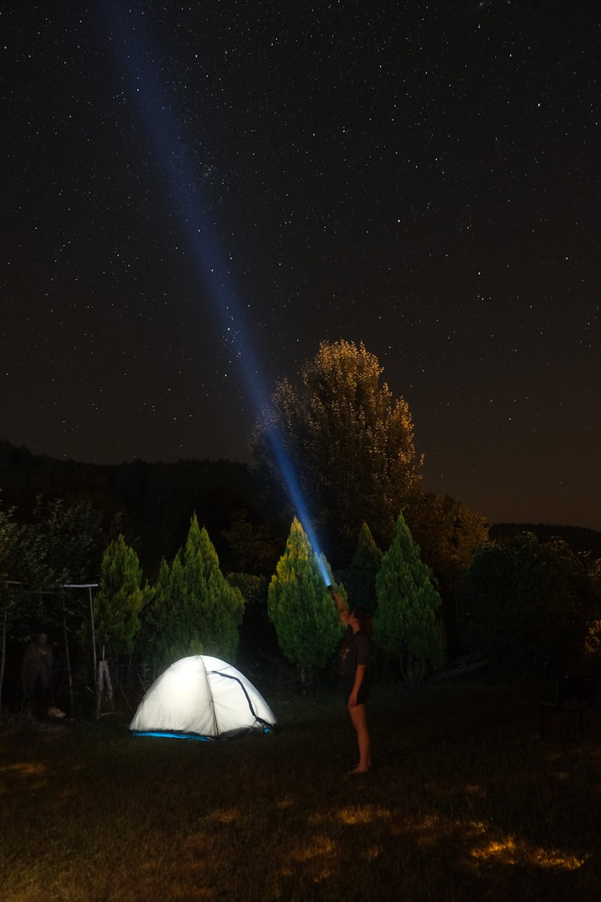 A person using a flashlight near a tent under a starry night sky, surrounded by trees.