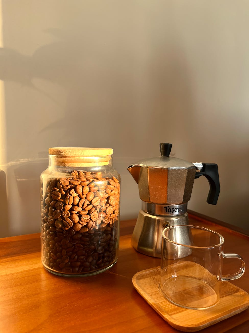 A cozy setup featuring a jar of coffee beans, Moka pot, and glass cup in warm lighting.
