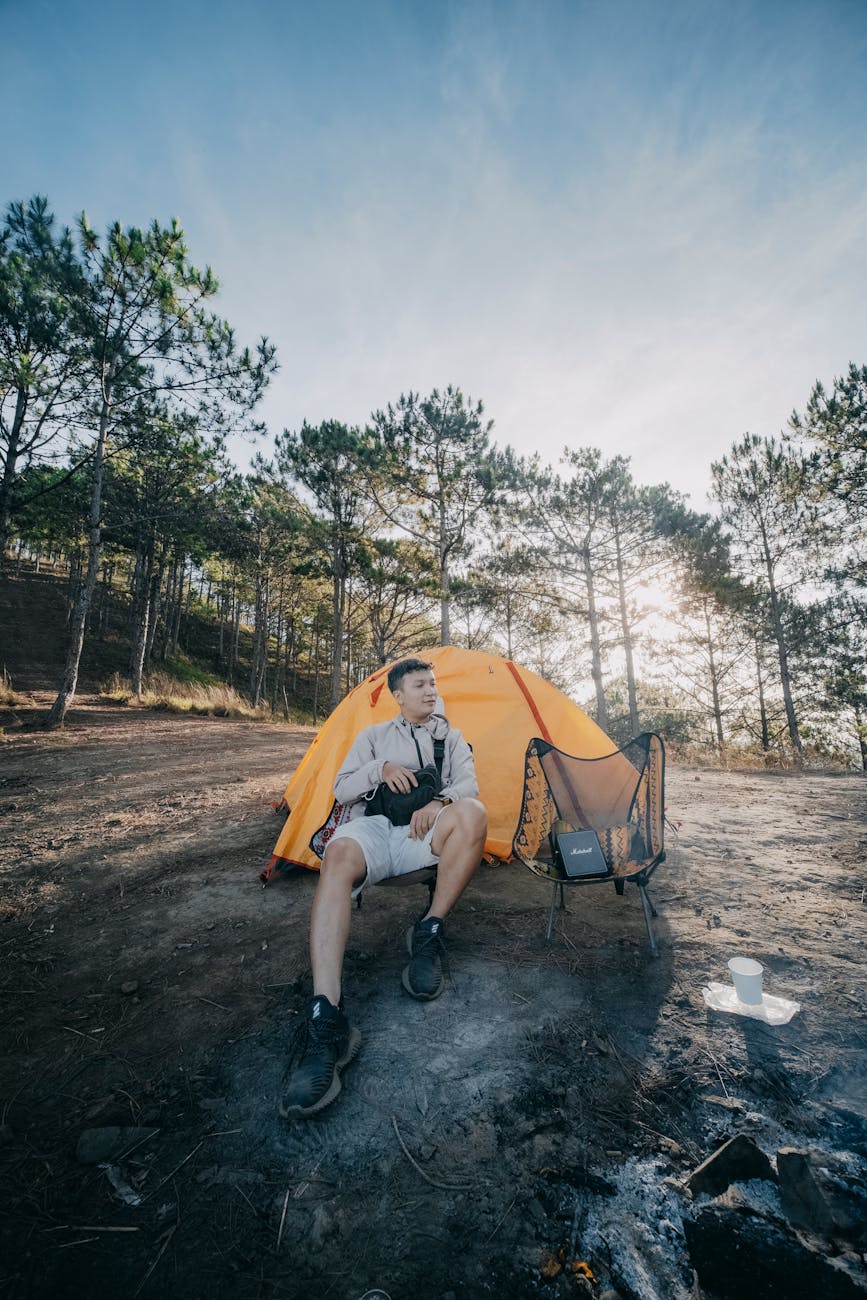 Young man sitting by a bright orange tent in a forest campsite enjoying nature.