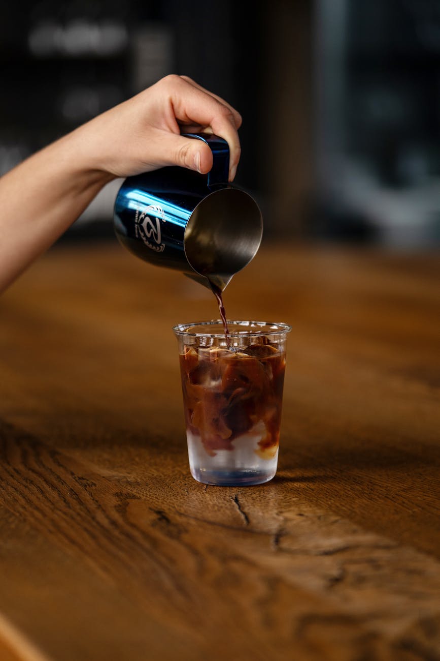 Hand pouring iced coffee from a pitcher into a glass on a wooden table. Close-up shot.