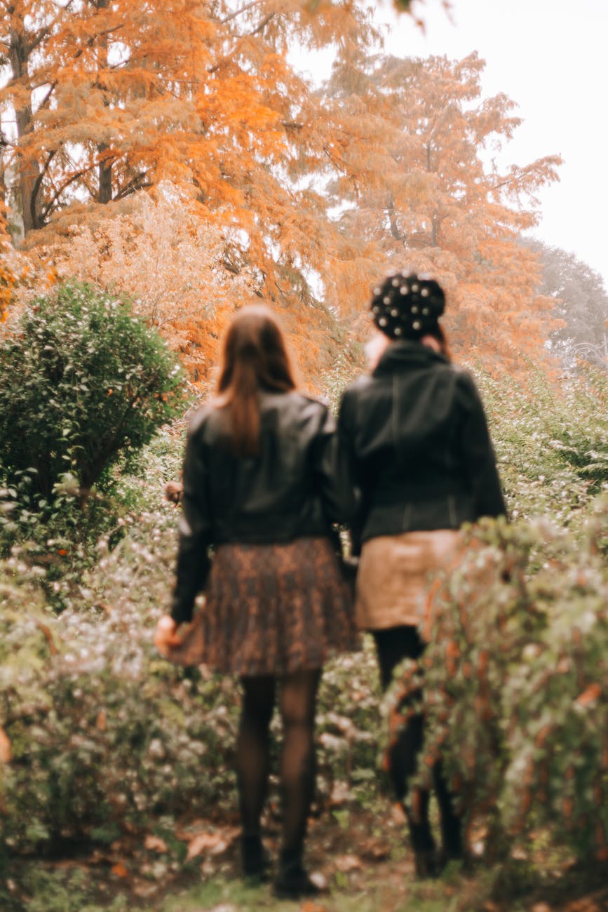Two women in leather jackets walk through a vibrant autumn forest, surrounded by fall foliage.