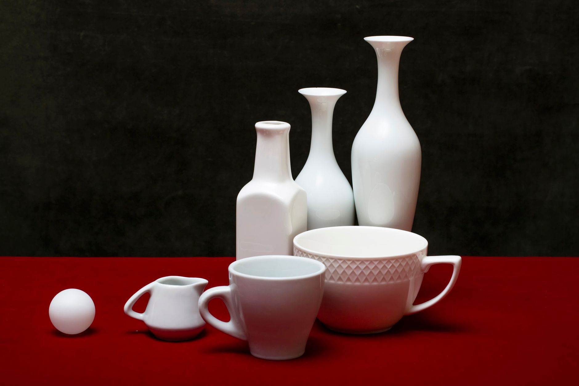 A collection of white ceramic vases and cups elegantly arranged on a vivid red surface against a dark backdrop.