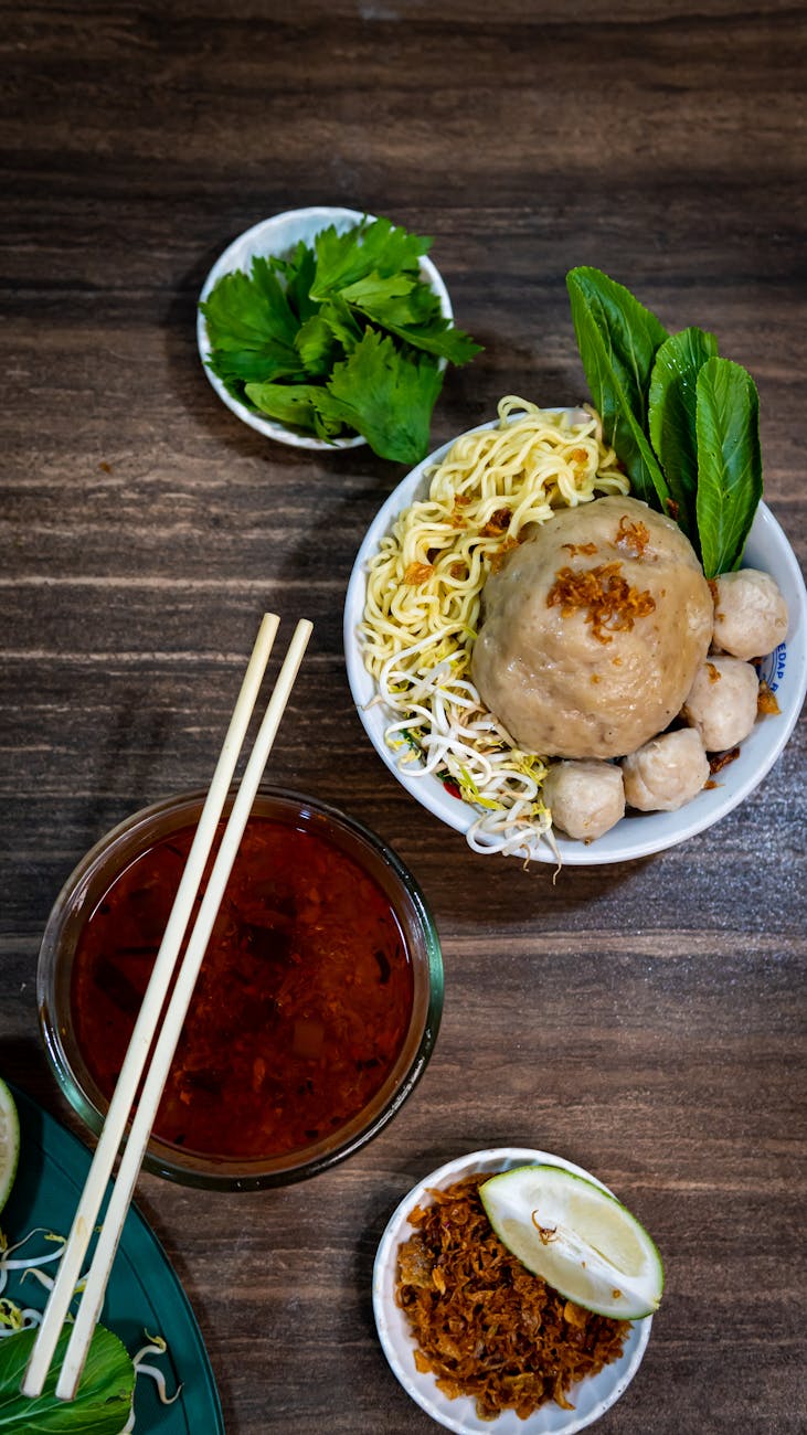 Flavorful Indonesian noodles with large meatball, sauce, and herbs on a wooden table.