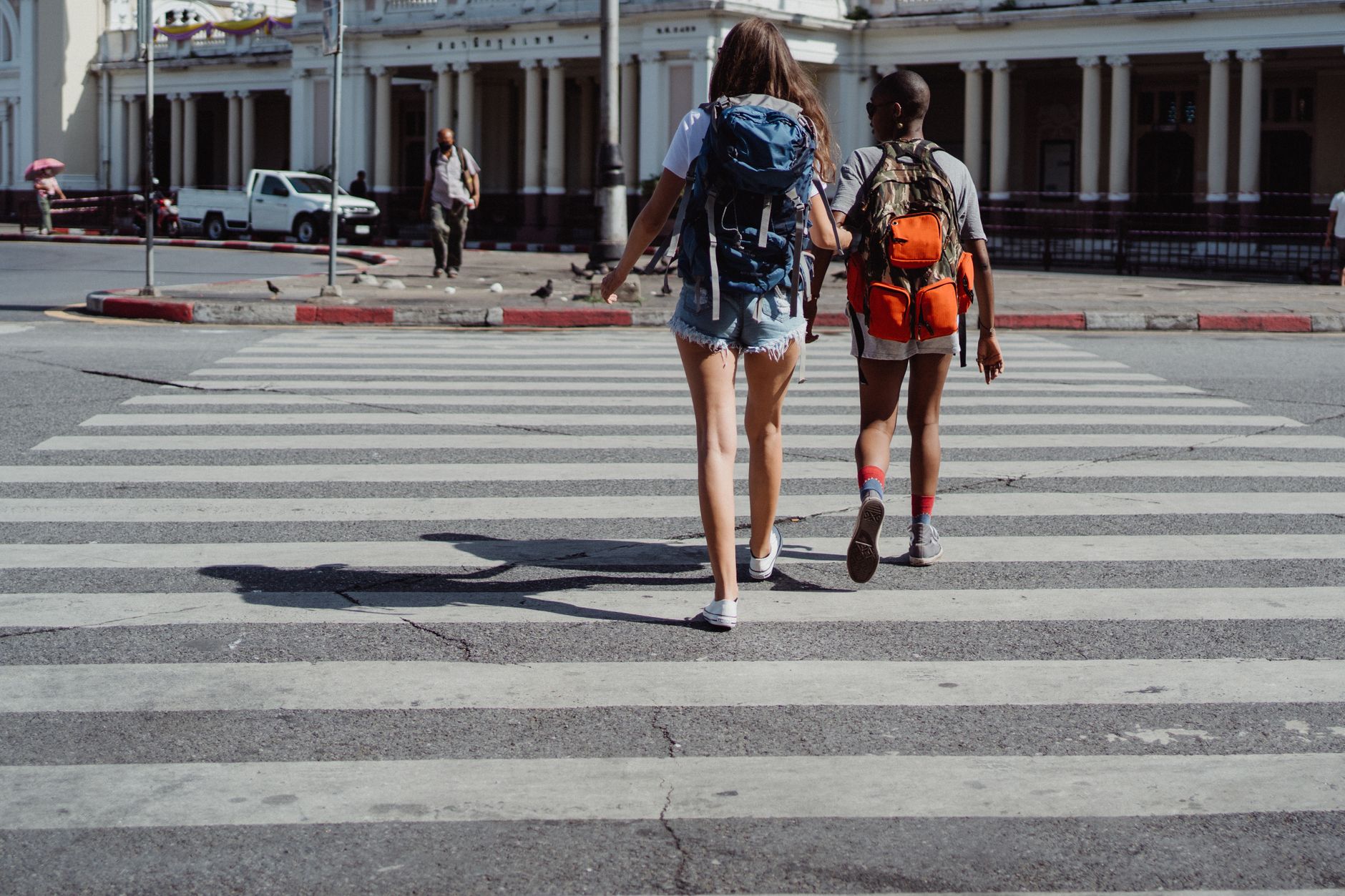 Two women with backpacks crossing a pedestrian lane in an urban setting on a sunny day.