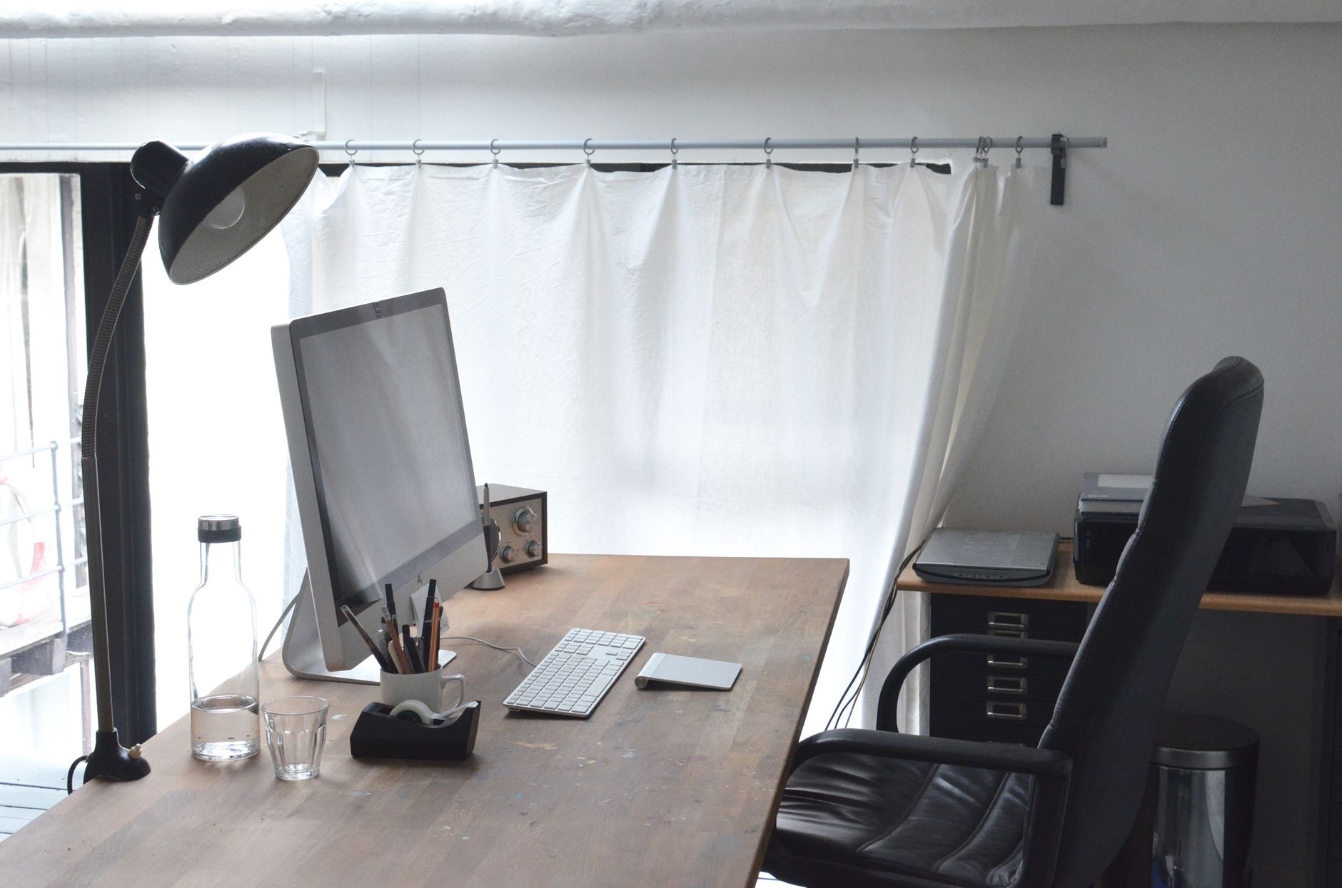 Modern computer with stationery and lamp arranged on wooden table near comfortable leather chair in office