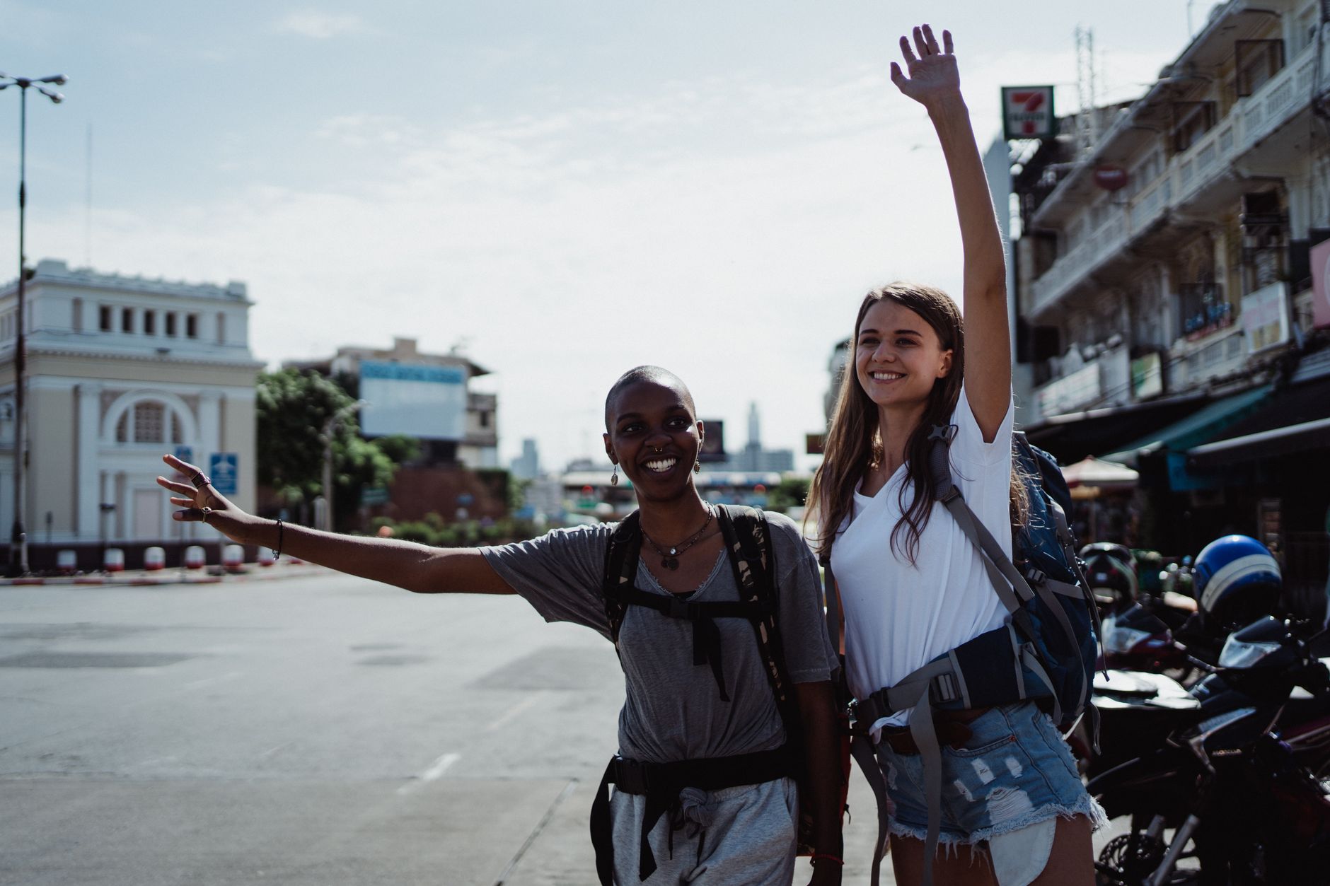 Two women with backpacks cheerfully hitchhiking on a sunny urban street.
