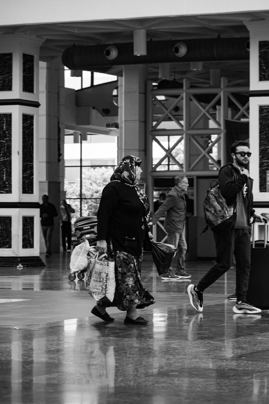 Black and white photo of people walking with luggage in a modern airport terminal.