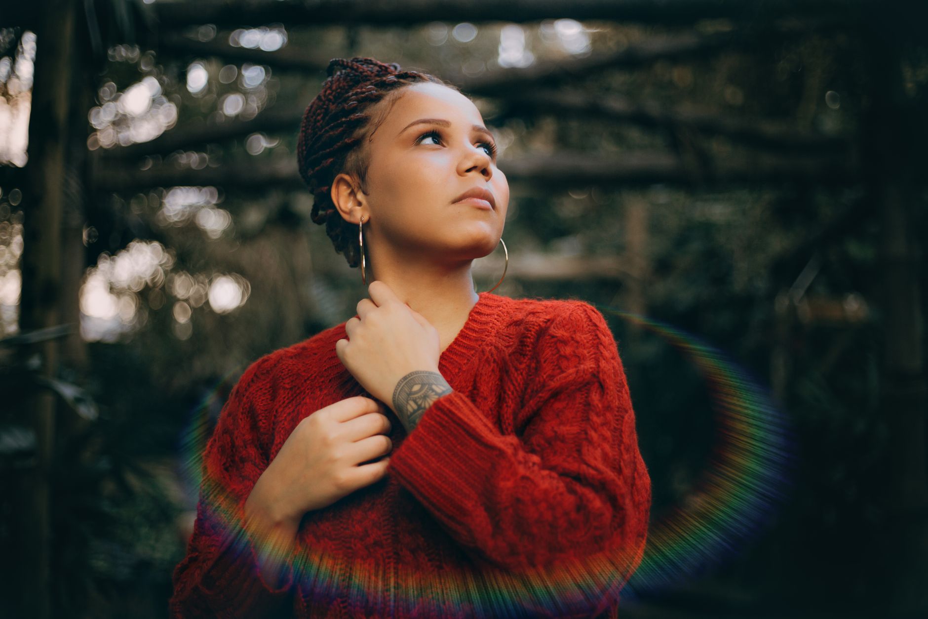 Portrait of a woman with braided hair in a red sweater, looking thoughtfully to the side outdoors.