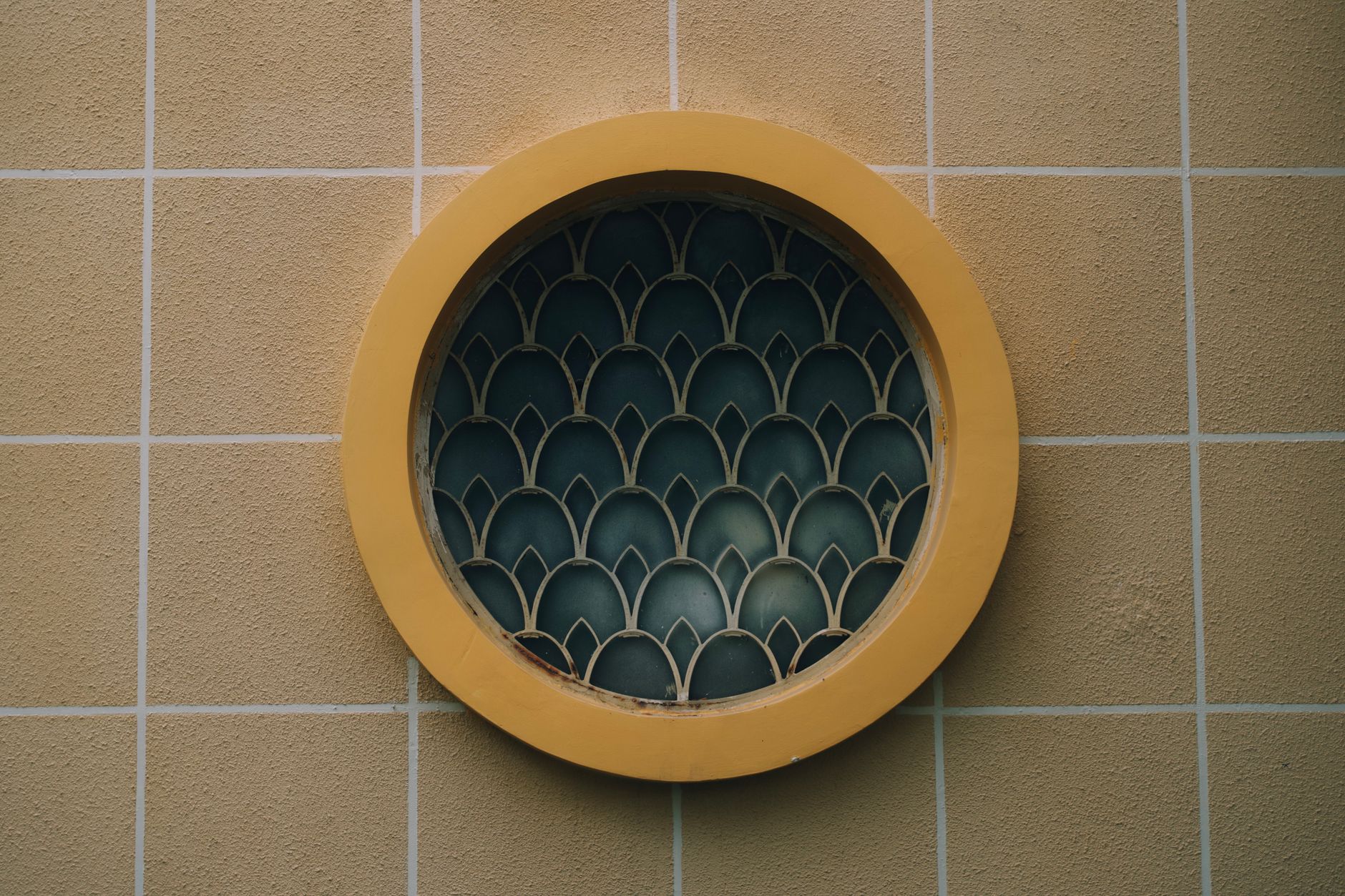 Circular window with decorative grille set in a yellow tiled wall, showcasing geometric patterns.