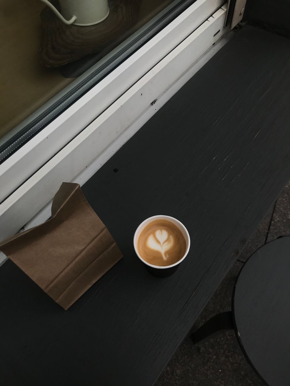 A cup of latte with heart art on black table outdoors, near brown paper bag.