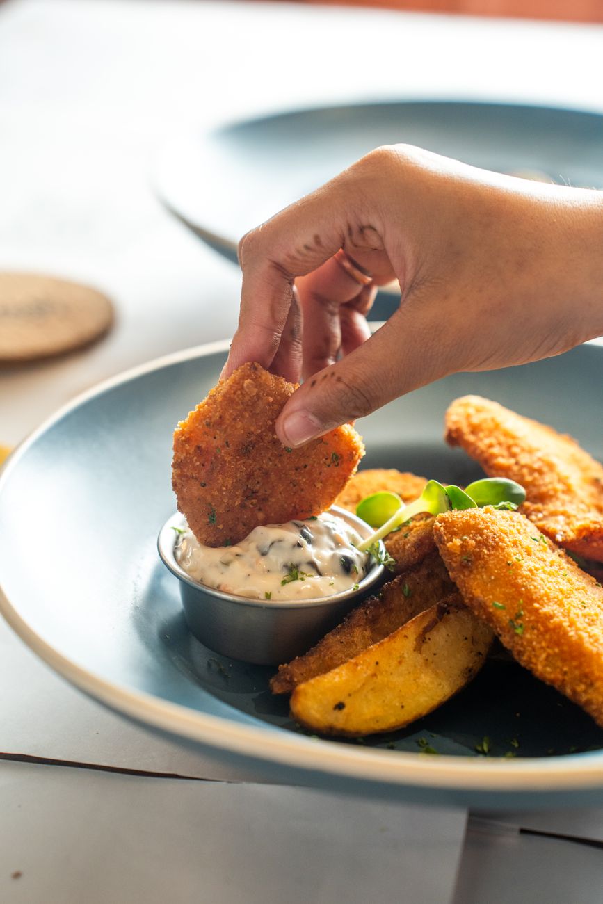 Hand dipping a crispy nugget into creamy sauce with potato wedges on a plate.