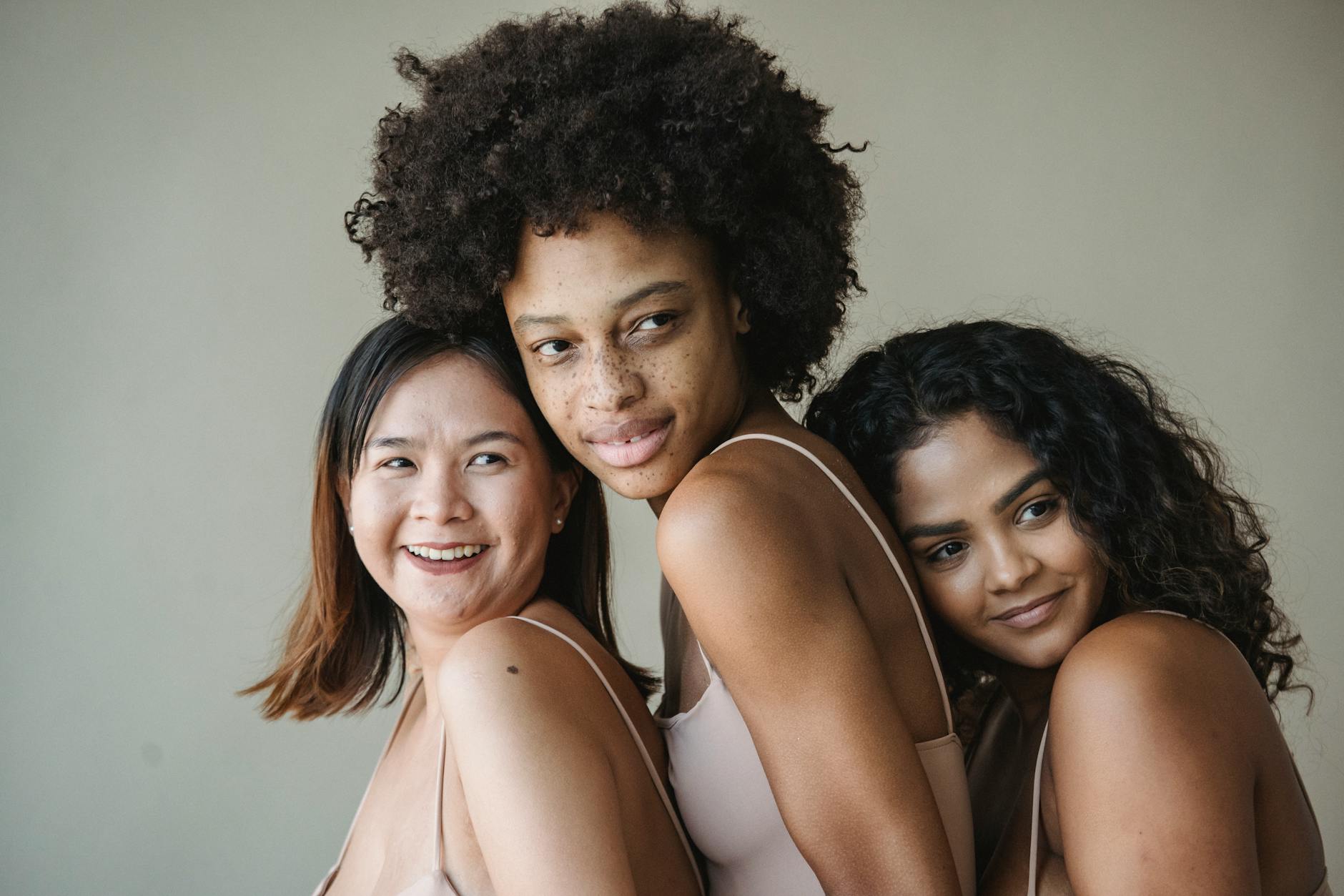Three diverse women embracing and smiling, showcasing beauty and friendship in a natural setting.