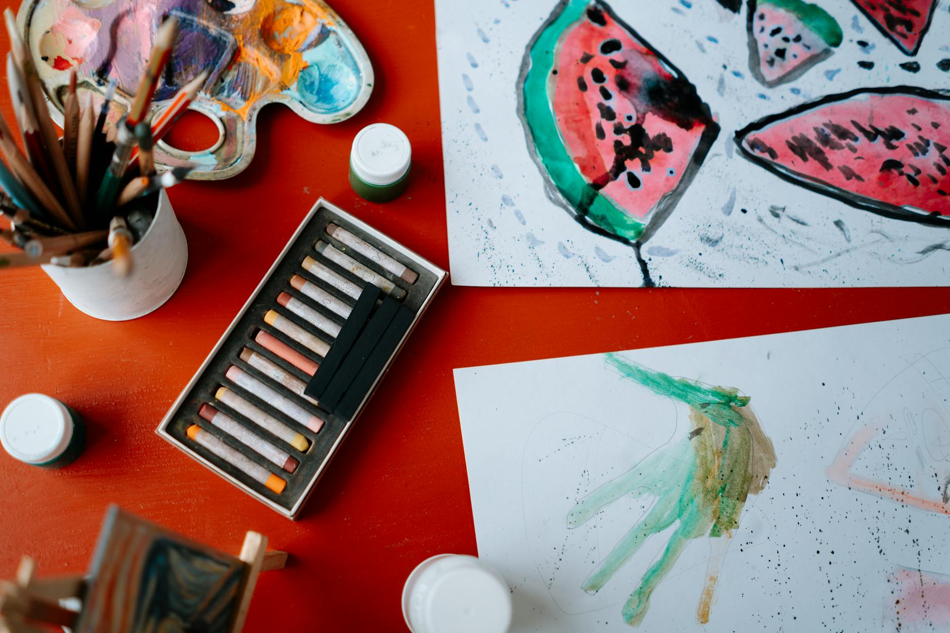 Creative art setup featuring watercolors and pastels on a red table, showcasing artistic expression.