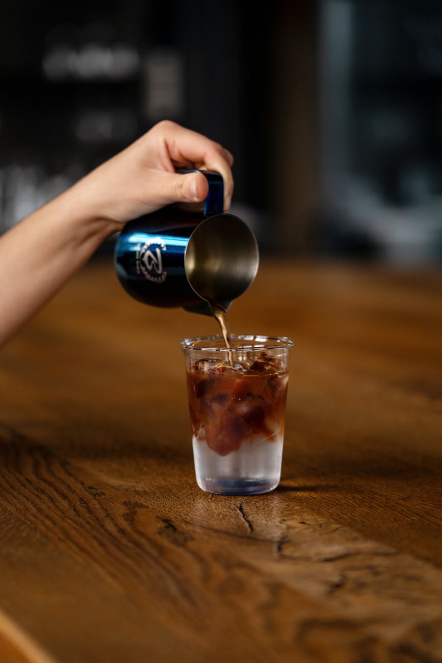 Hand pouring coffee from a metallic pitcher into a glass on a wooden surface, captured with a selective focus.