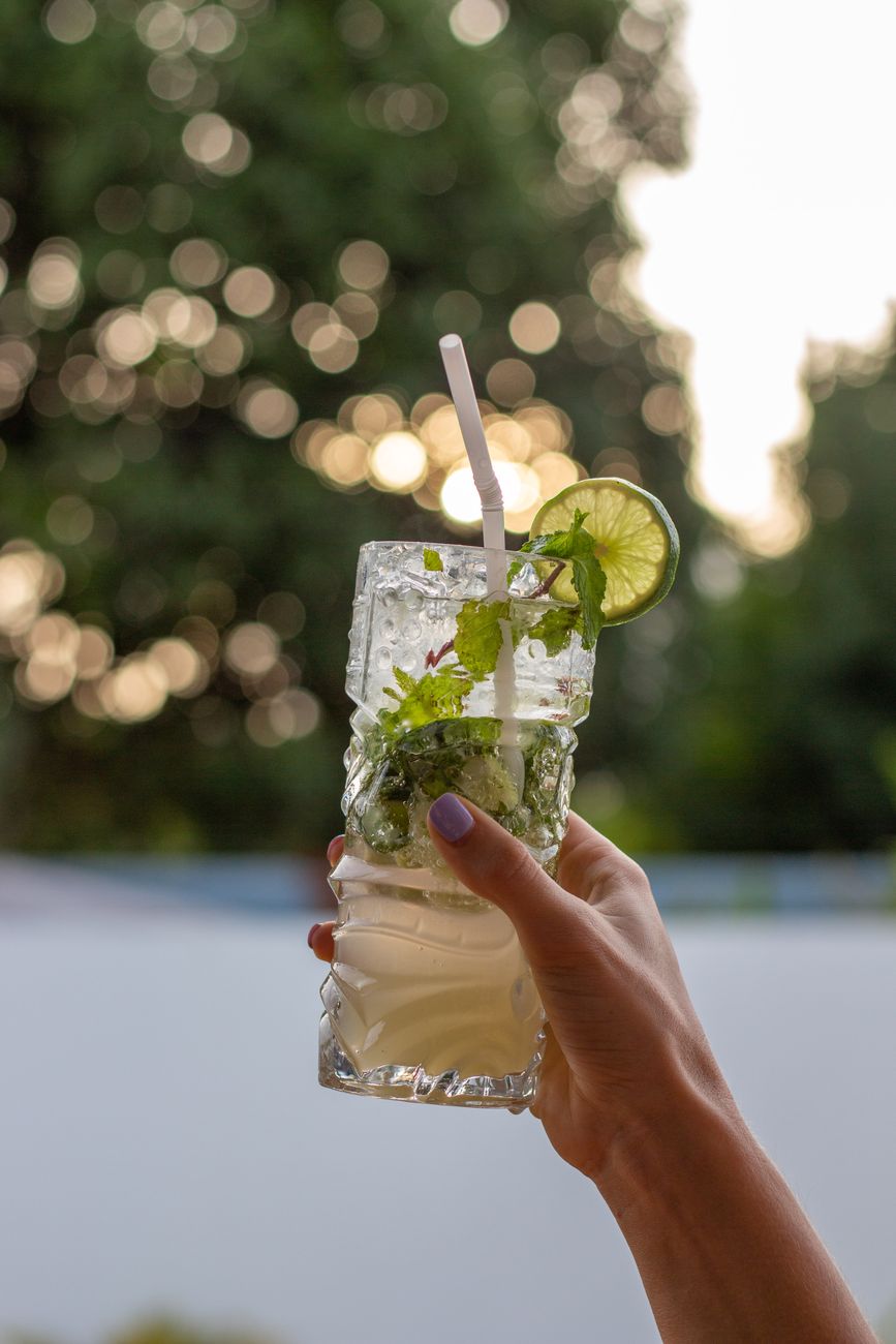 Hand holding a refreshing lime mojito cocktail with mint, ice, and straw, against a bokeh background.