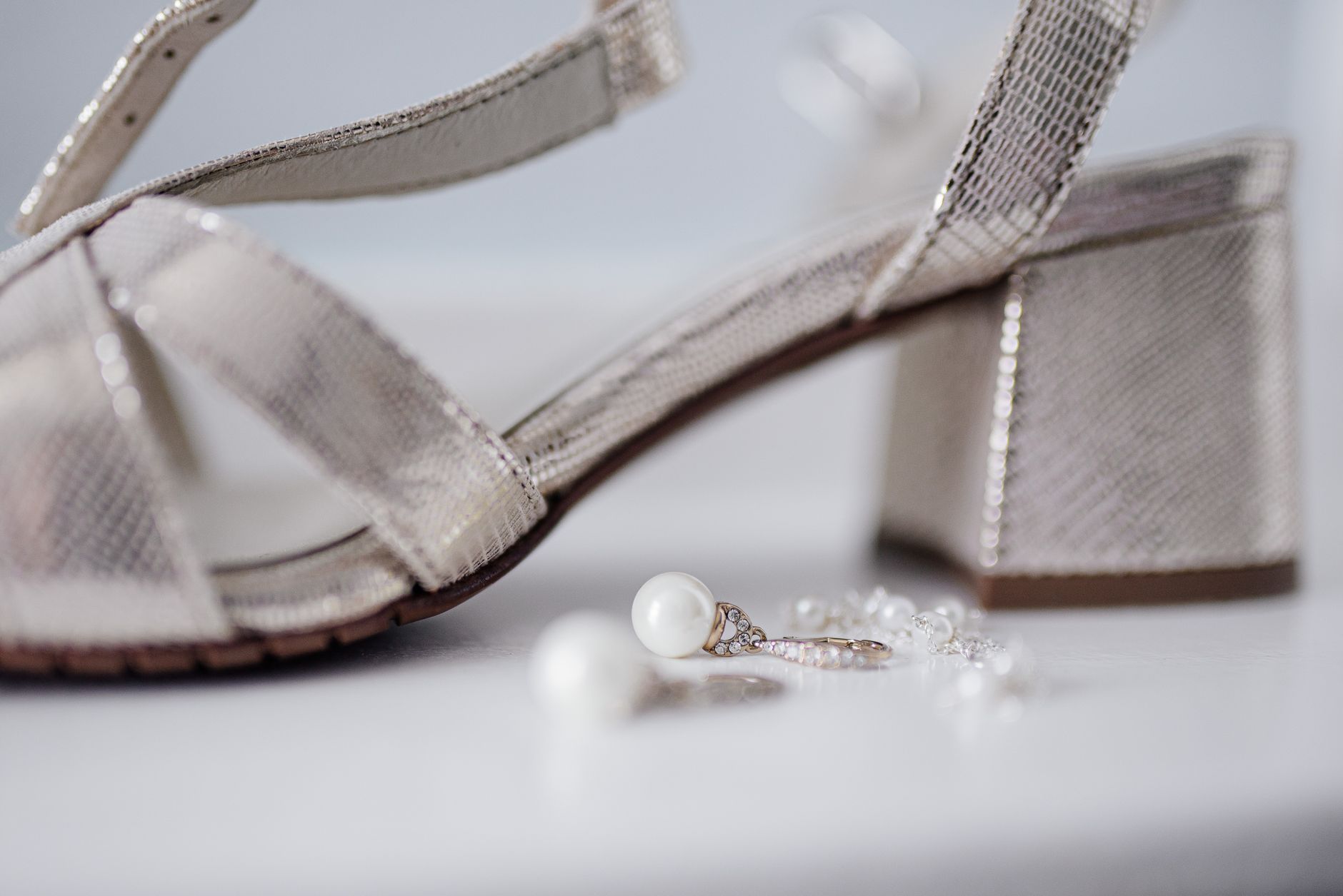 Close-up of bridal block heel sandals paired with pearl earrings on a white surface.