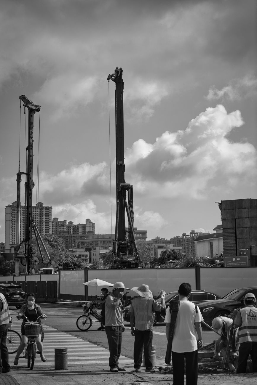 Black and white image of construction workers at a city site with cranes and pedestrians.