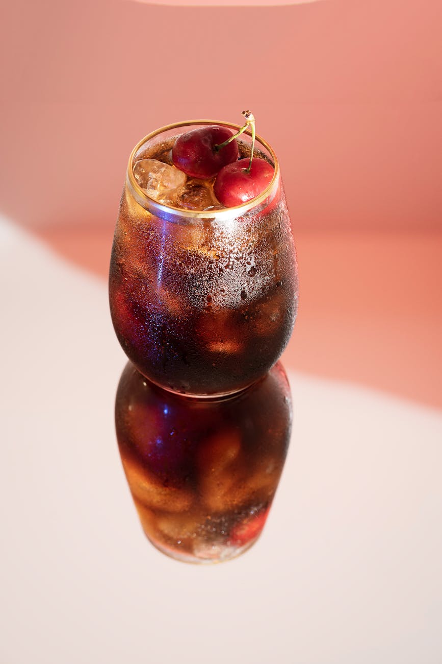 A cold glass of cherry cola with ice, featuring a vivid reflection and cherries on top.