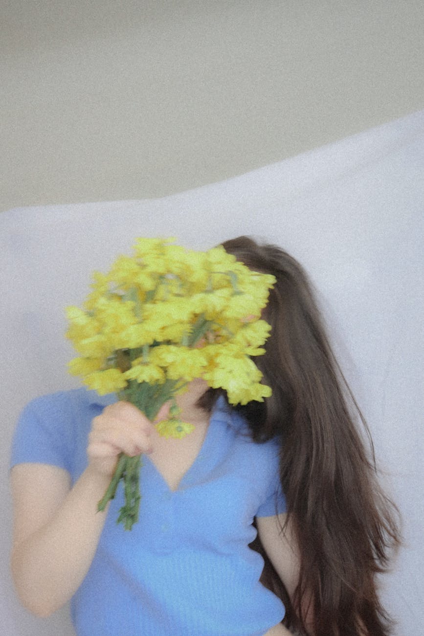 A woman in a blue shirt holds a bouquet of yellow flowers covering her face in a studio setting.