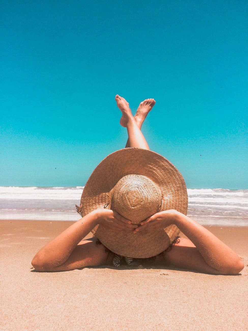 A woman in a straw hat relaxes on a sunny beach, enjoying the clear blue sky and ocean waves.